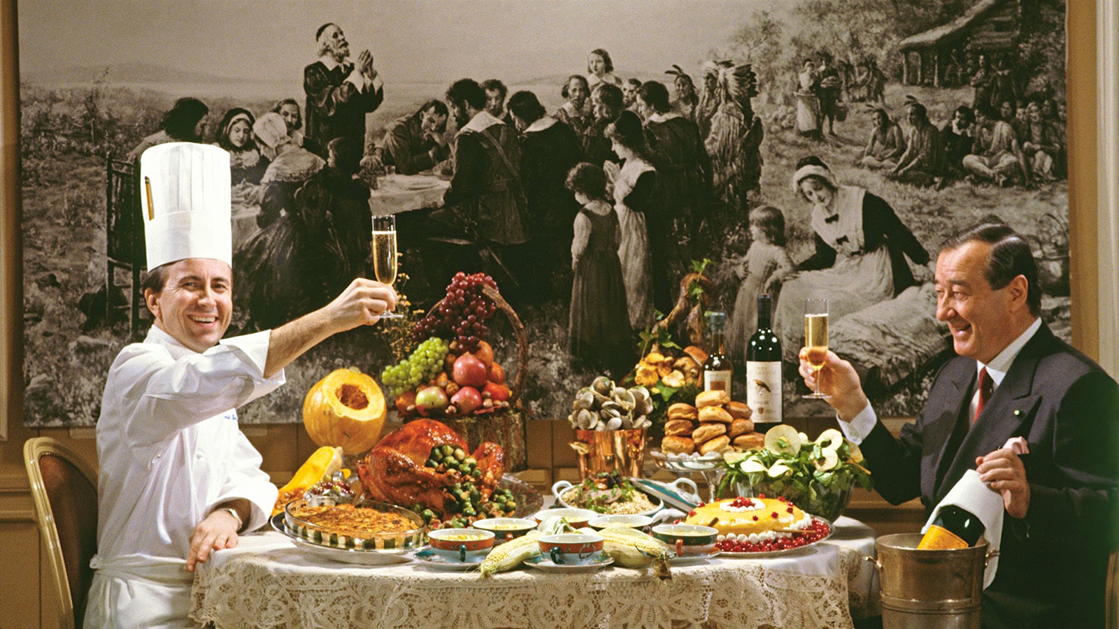 Chef Daniel Boulud holding up a glass of sparkling wine at a laden table at Le Cirque restaurant